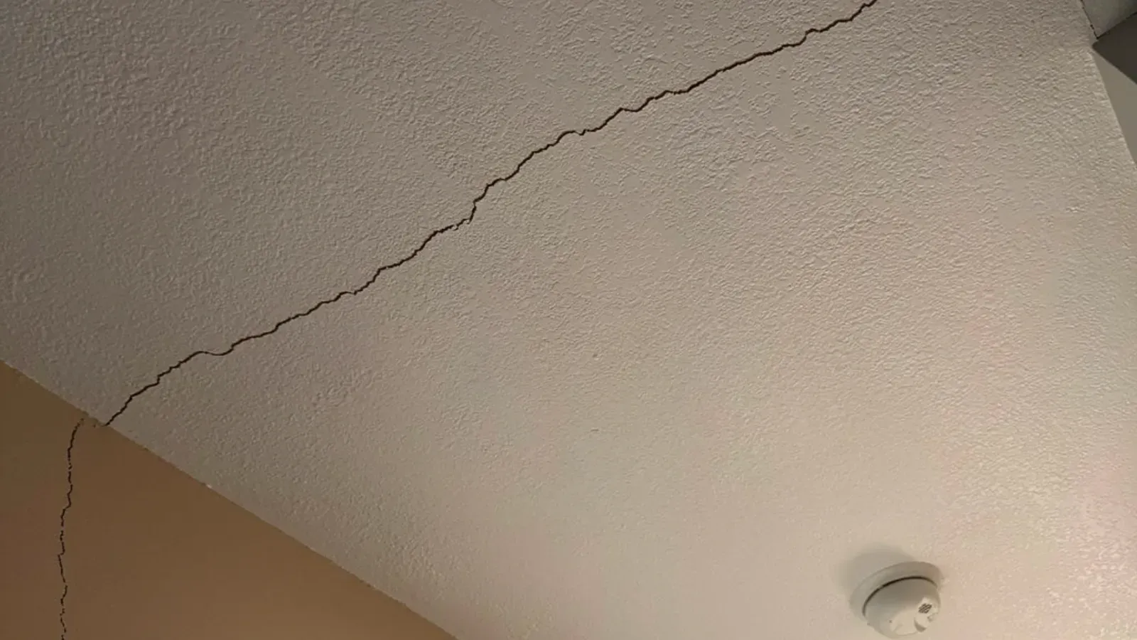 A long crack running diagonally across a lightly textured painted ceiling toward the corner where the ceiling meets the wall, with a round white smoke detector mounted nearby