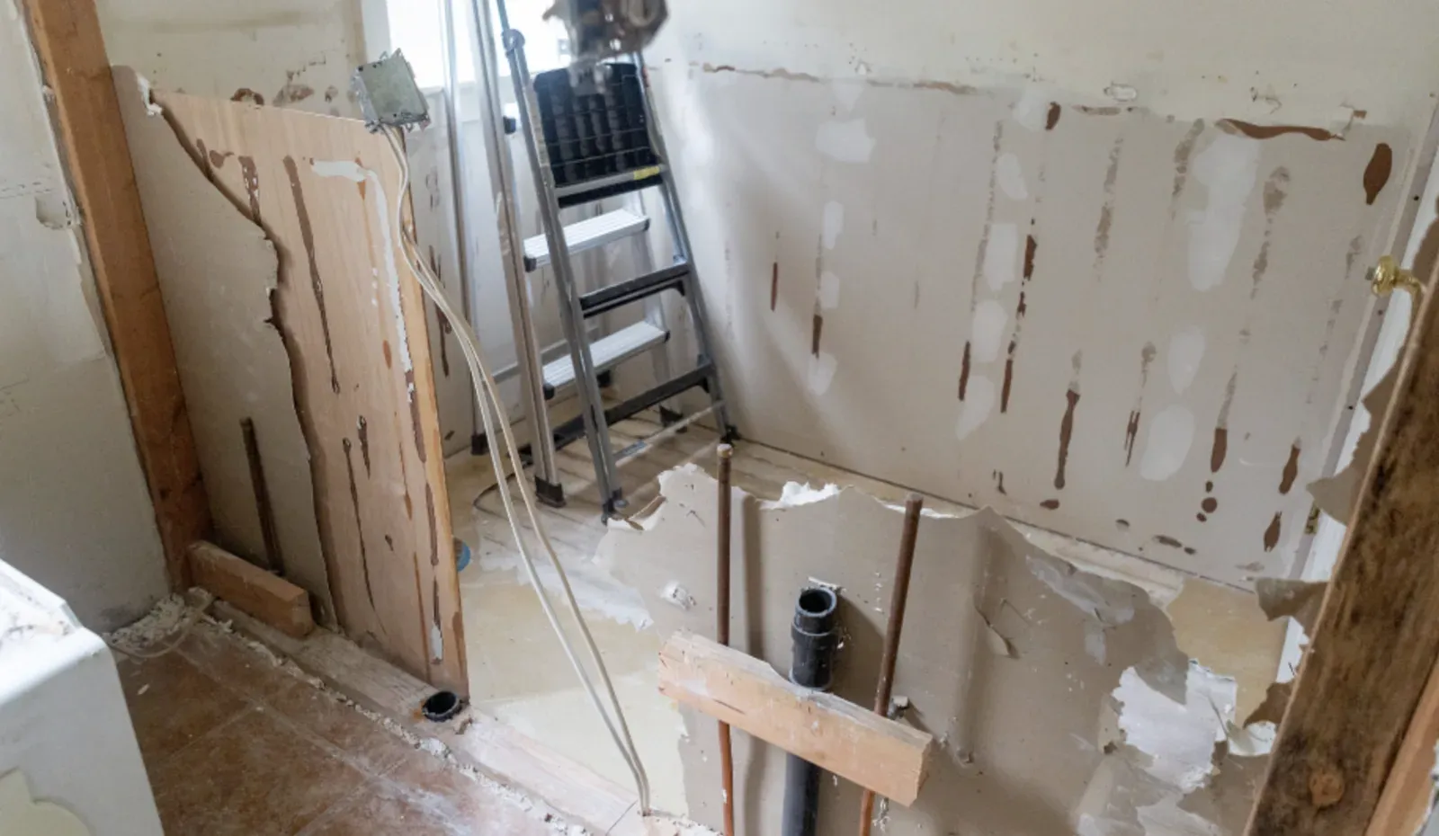 Bathroom mid-demolition with drywall torn back to the studs, exposed copper and PVC plumbing rough-in, a removed drywall panel leaning against the wall, and a step ladder