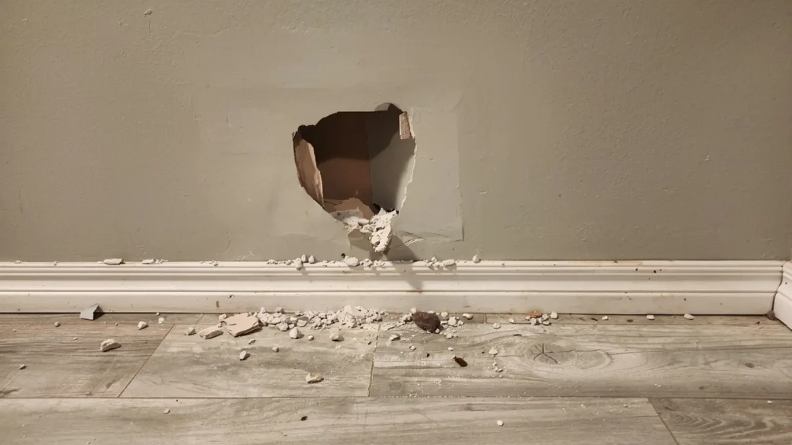 Broken hole low on an interior drywall wall near a corner, gypsum debris scattered on the floor along the baseboard