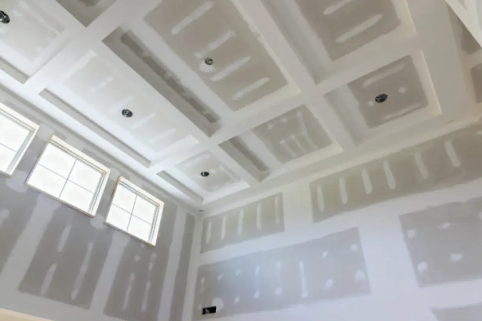 Tall room with a coffered drywall ceiling and clerestory windows, seams taped and fasteners spotted throughout before sanding