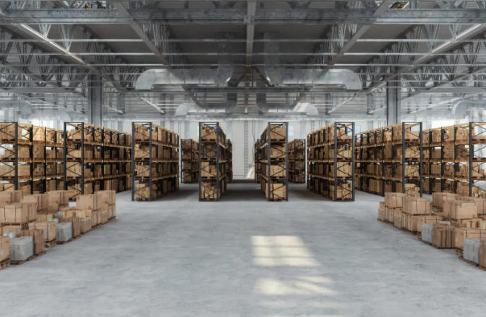 Wide warehouse floor with shelving aisles of stacked cartons under an exposed steel roof truss system, daylight falling across the concrete slab